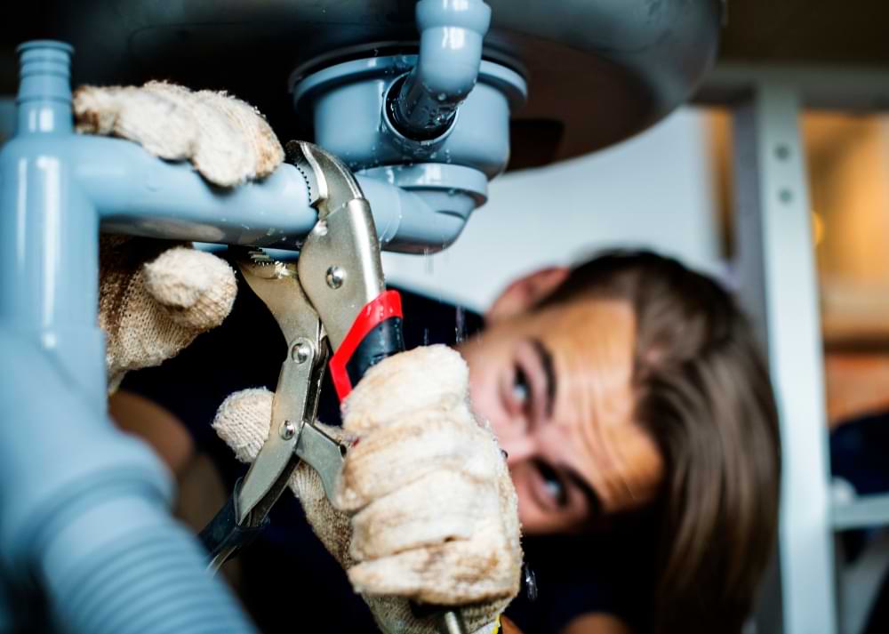 A plumber wearing gloves tightens a pipe under a sink using a wrench.