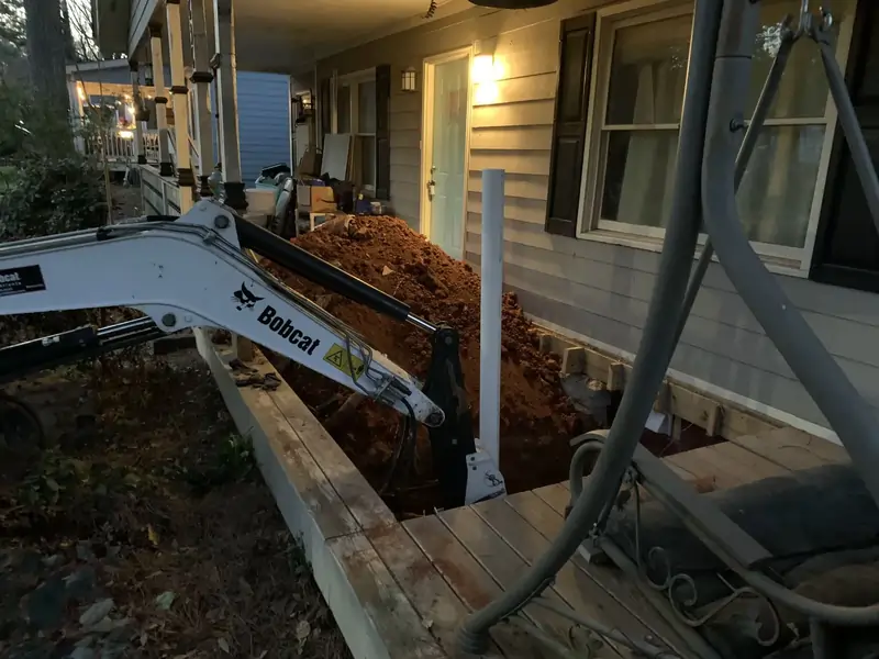 A small excavator works near a house, digging a trench on a porch with exposed soil piled up.