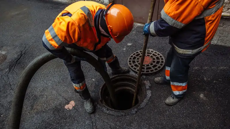 Two workers in orange safety gear and helmets clean a sewer, inserting a hose and rod into a manhole on a paved street.