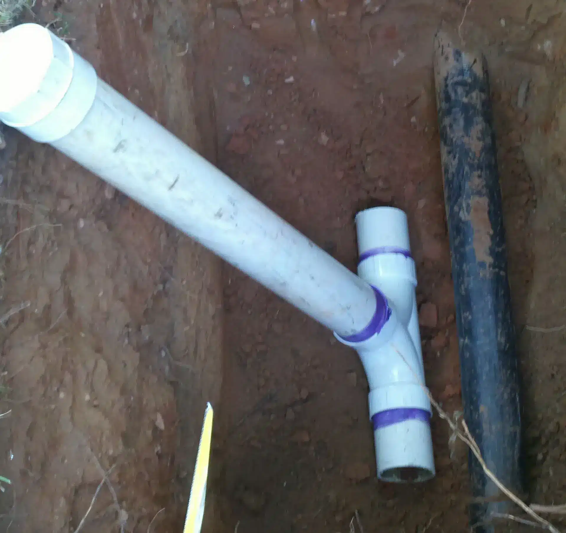 A white PVC plumbing pipe with purple connectors is fitted into a dirt trench, alongside a black pipe.