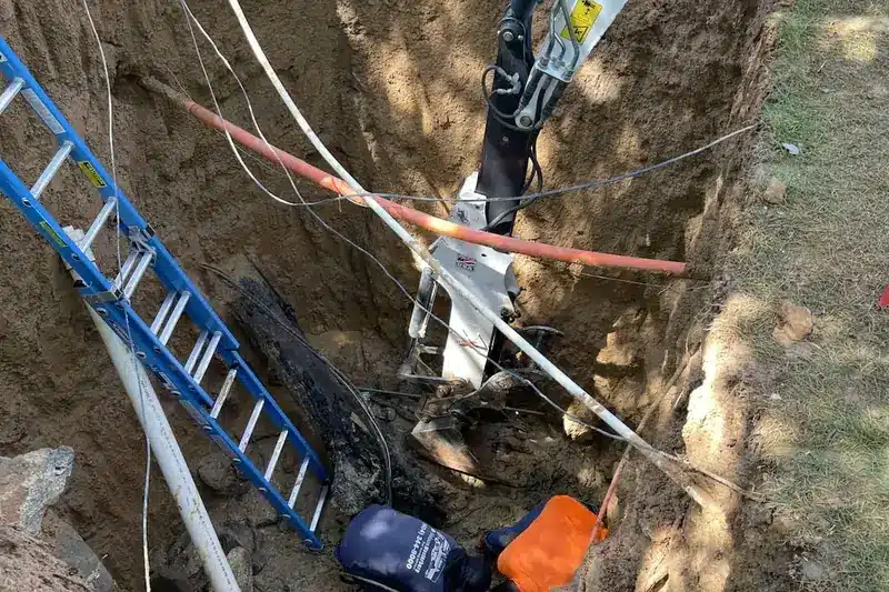A deep trench with a blue ladder on the left, orange and white utility pipes crossing above.