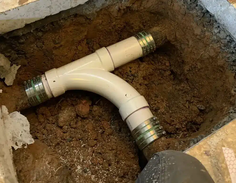 A pipe junction with metal clamps is visible in an excavated dirt area, showing plumbing installation work in progress.