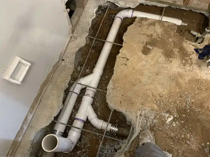 A construction site shows exposed plumbing with PVC pipes beneath a concrete floor, surrounded by dirt.