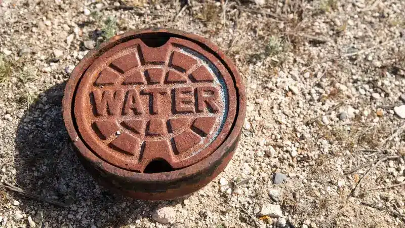 Rusty water valve cover with a bold "WATER" inscription, set in sandy terrain with sparse grass, conveying an aged, rugged outdoor setting.