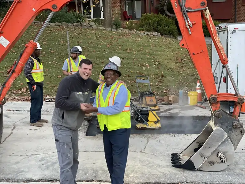 Two men wearing construction attire smile and shake hands at a worksite between two excavators, with other workers visible in the background.