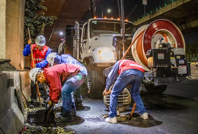 Workers in hard hats and reflective vests clear a storm drain at night.