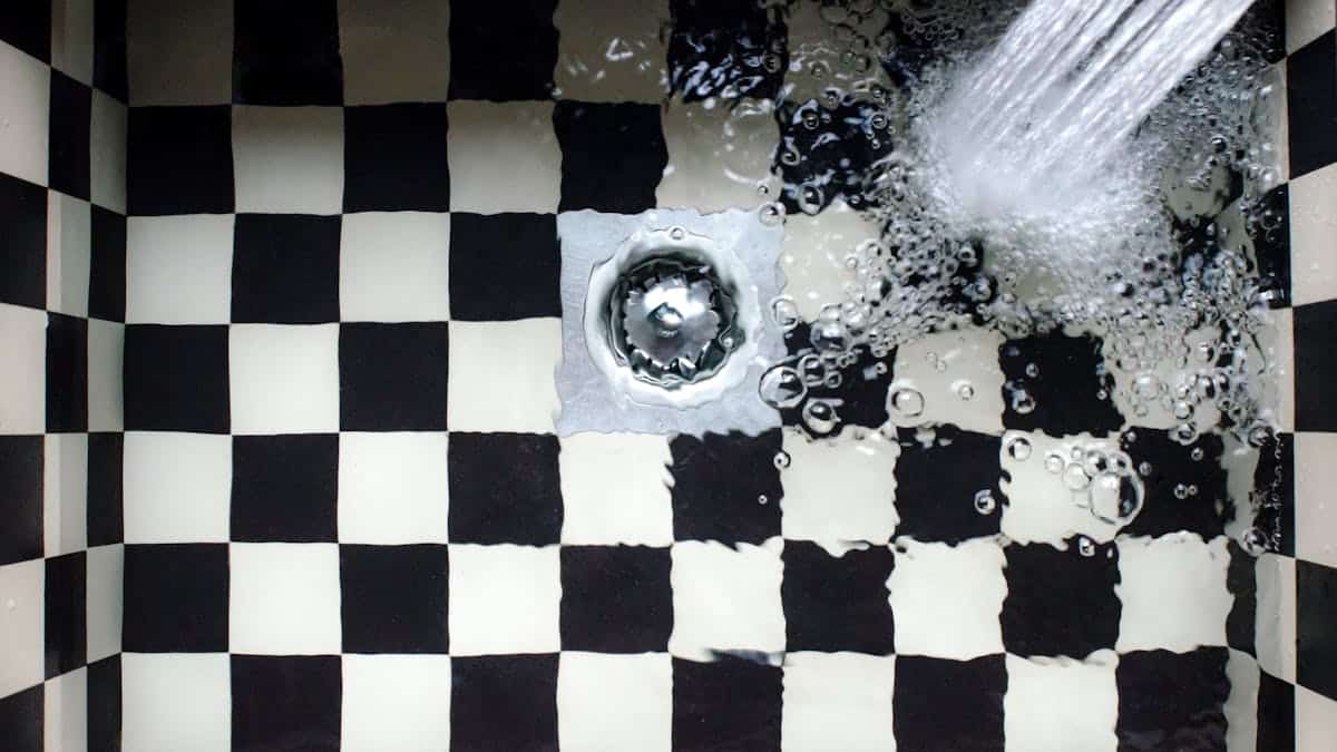 Water pouring down a shower or bathtub drain surrounded by black and white checkered tiles.