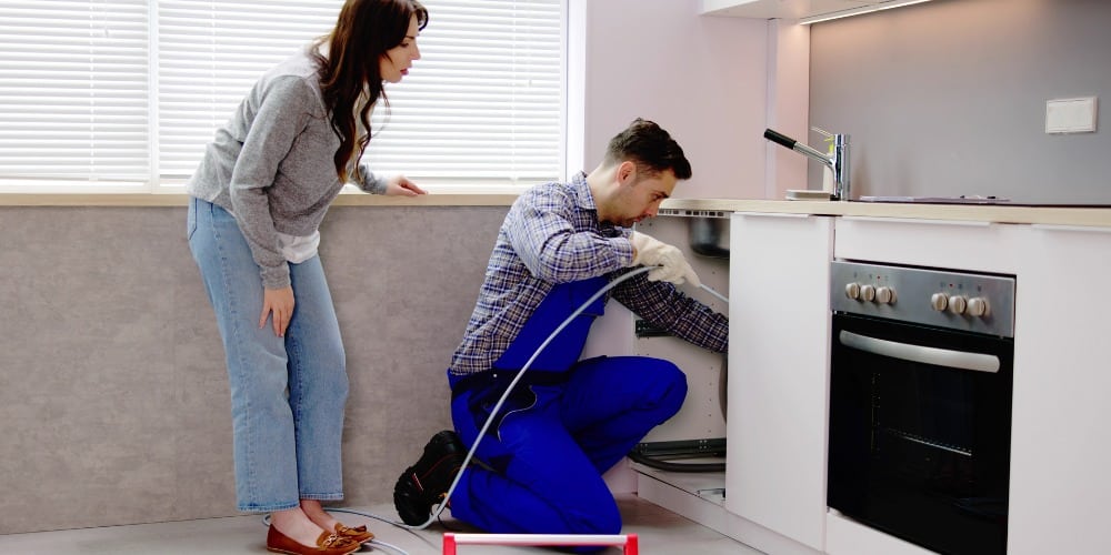A plumber in blue overalls uses a drain snake to fix a kitchen sink
